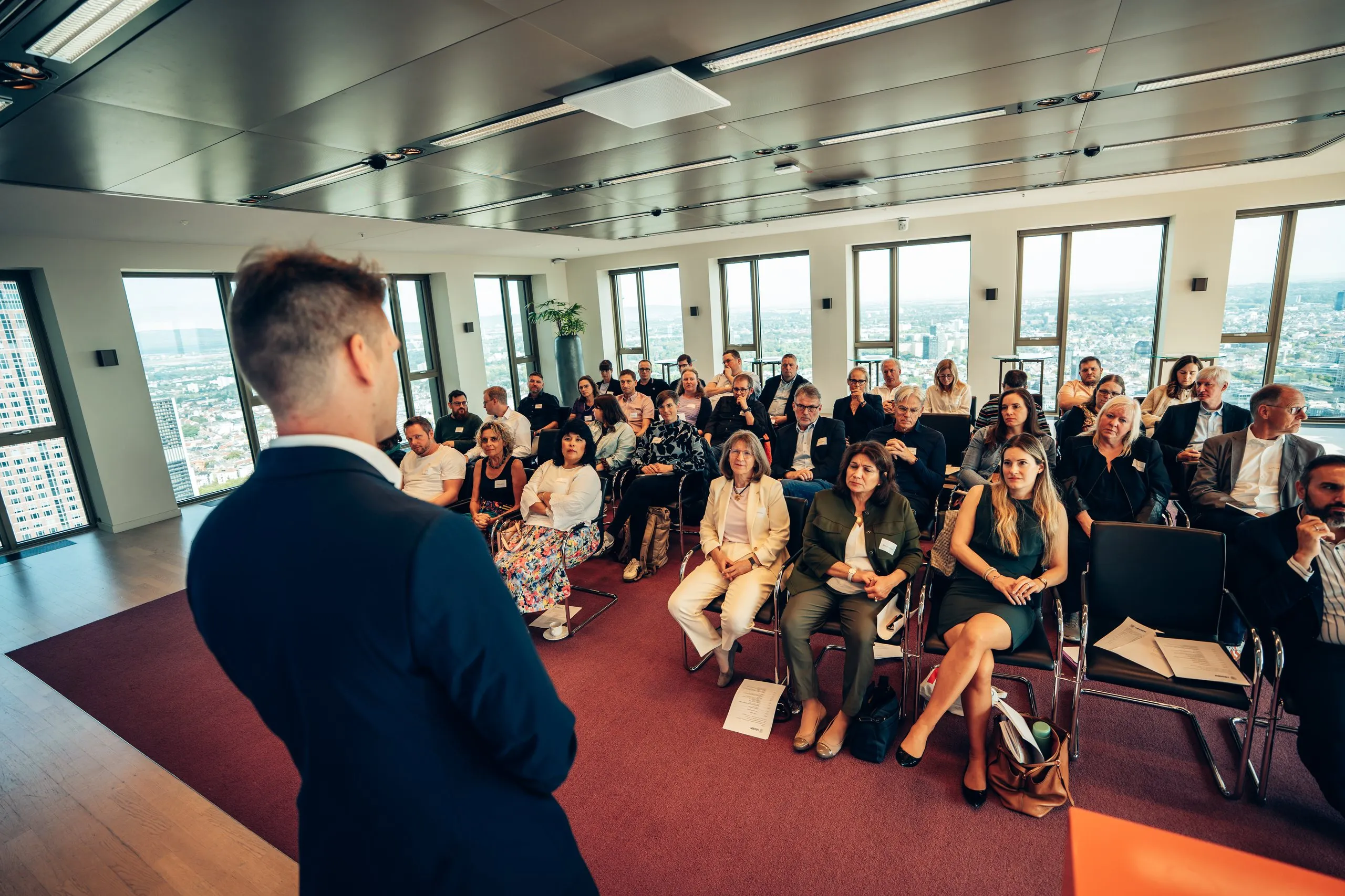 Pascal Kayser bei einem Vortrag vor vollen Stuhlreihen, durch die Fenster der Blick über die Stadt